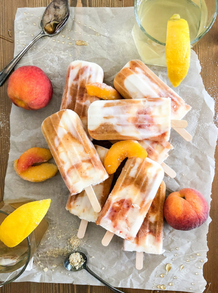 A pile of peach and greek yogurt popsicles with pieces of fresh peach around it. The popsicles are sitting on top of a piece of wax paper. 