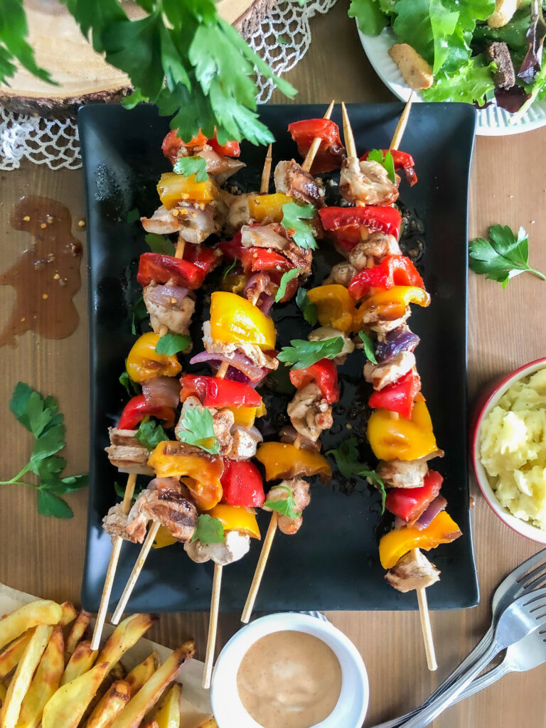 flat lay image of prepared marinated chicken and vegetable skewers on a black serving plate. The skewers are drizzled with a spicy red pepper sauce and topped with fresh parsley. 